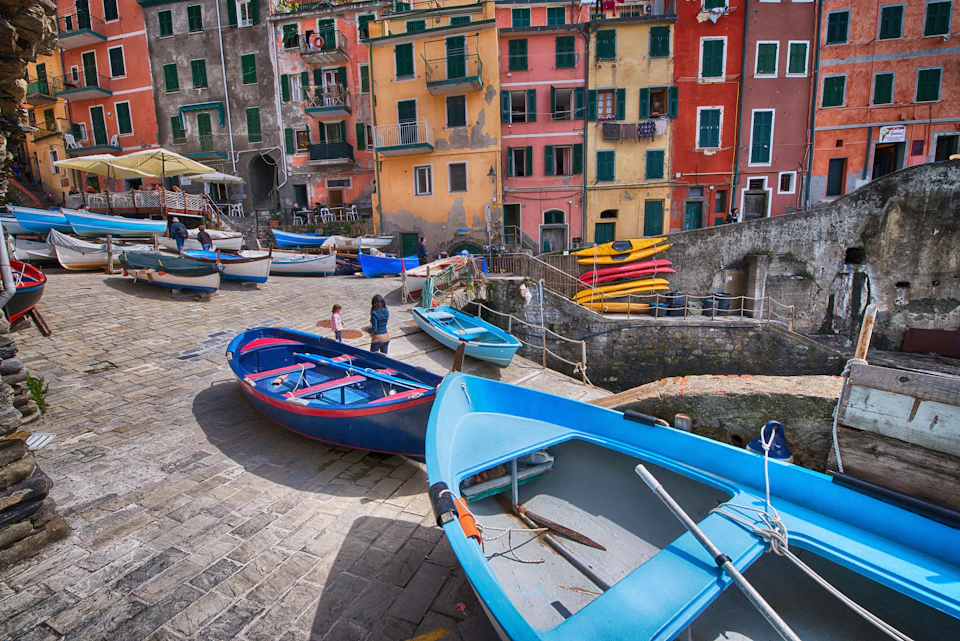 Colourful boats are parked on a stone dock with people walking nearby. Vibrant multicoloured buildings line the background.