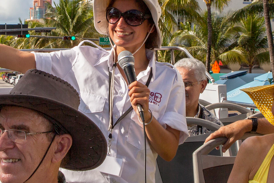 A woman tour guide with a microphone speaks to a group of tourists seated in an open-top bus, surrounded by palm trees and buildings.