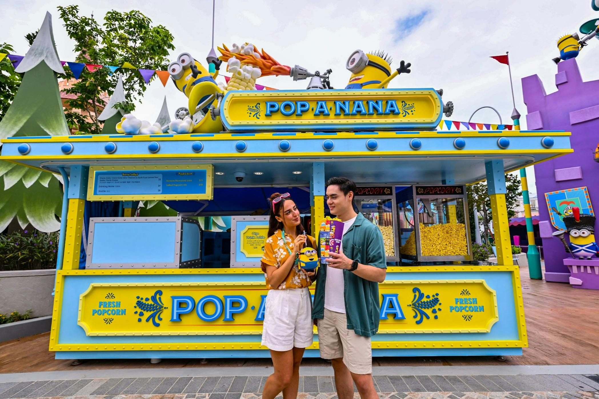 Duas pessoas em frente a uma banca de pipocas amarela e azul com personagens de desenhos animados, segurando uma caixa de pipocas.