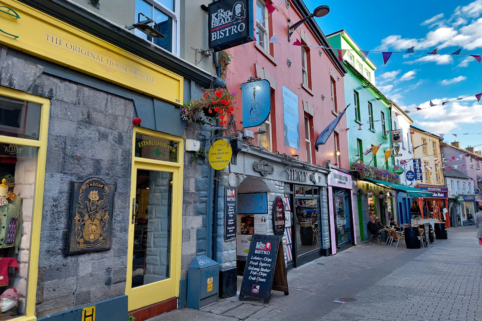 A colorful street lined with shops and bistros, featuring hanging flower baskets and signage under a bright blue sky.