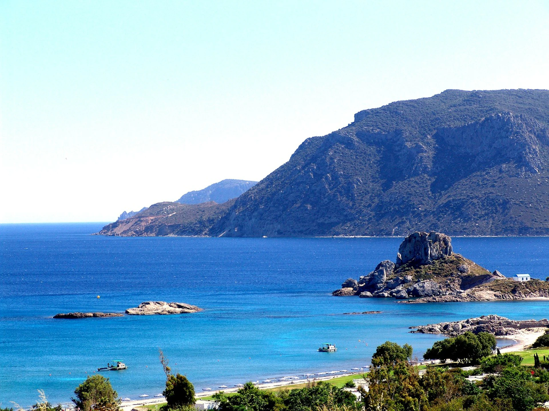 A coastal landscape with blue water, rocky hills in the background, small islands, and trees in the foreground.