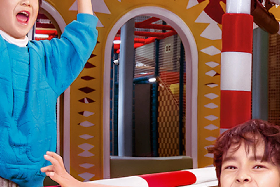 Children playing joyfully on a slide in a colorful indoor playground with a ball pit and decorative elements.