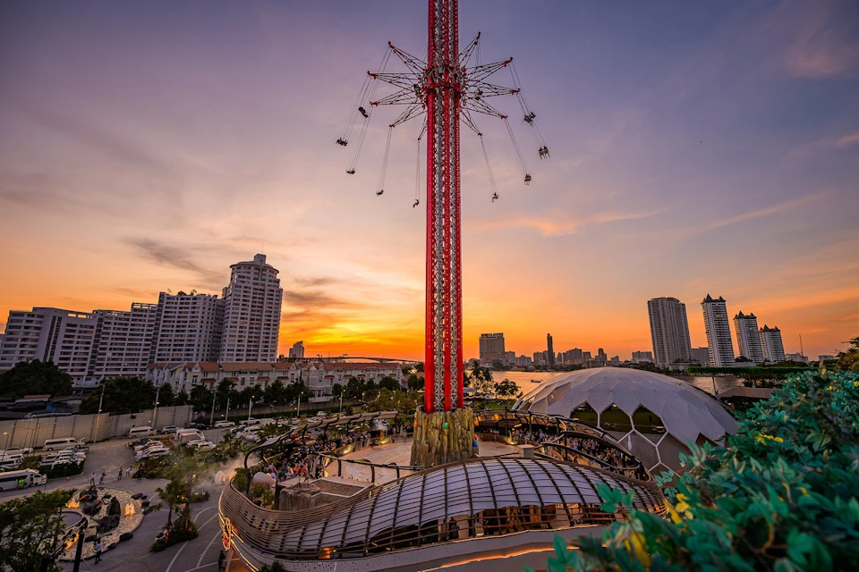 A tall amusement park ride with swinging seats at sunset, surrounded by buildings and greenery.