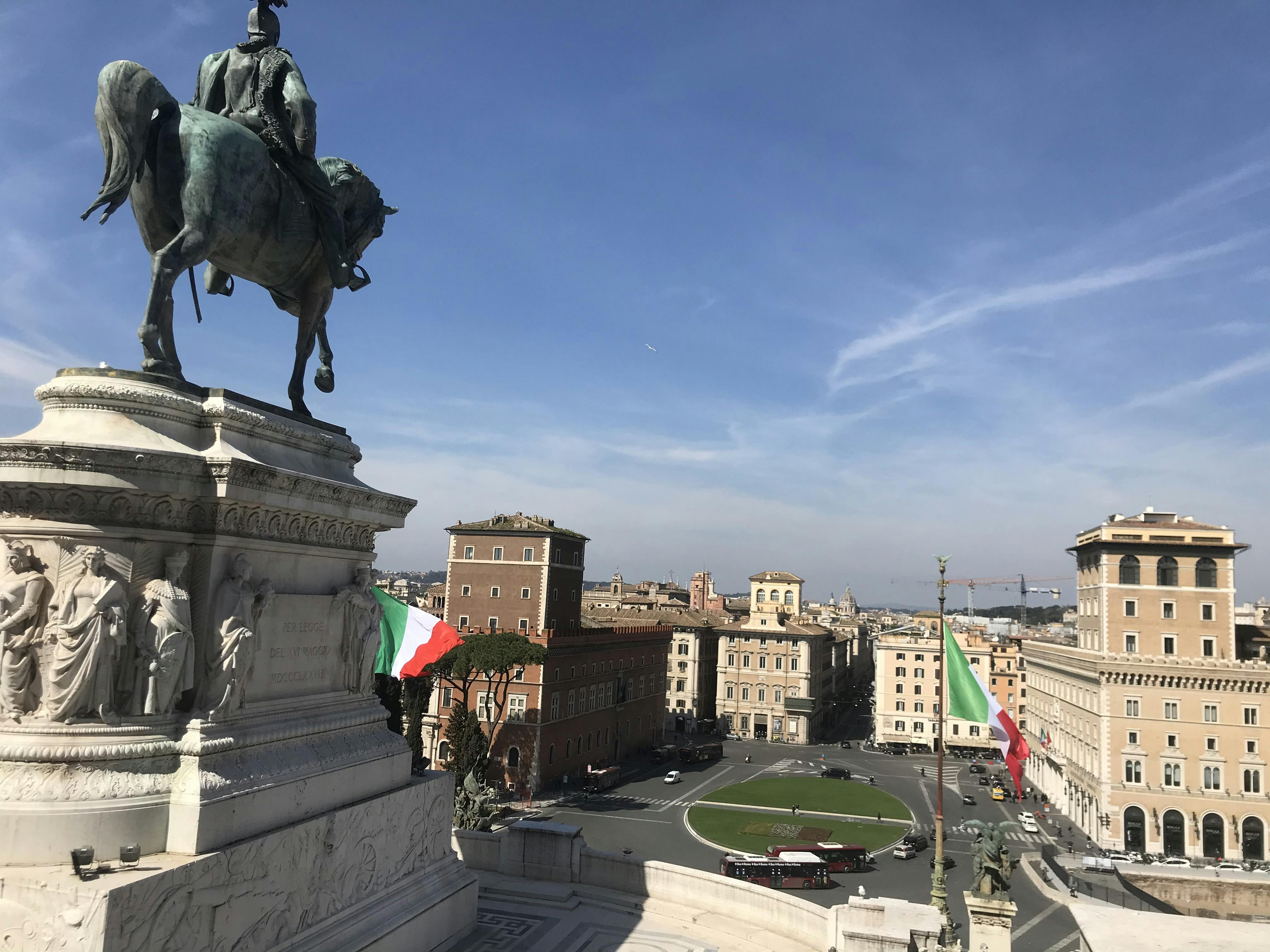 Statua equestre con bandiere italiane, che si affaccia su una piazza cittadina con edifici storici sotto un cielo azzurro e limpido.
