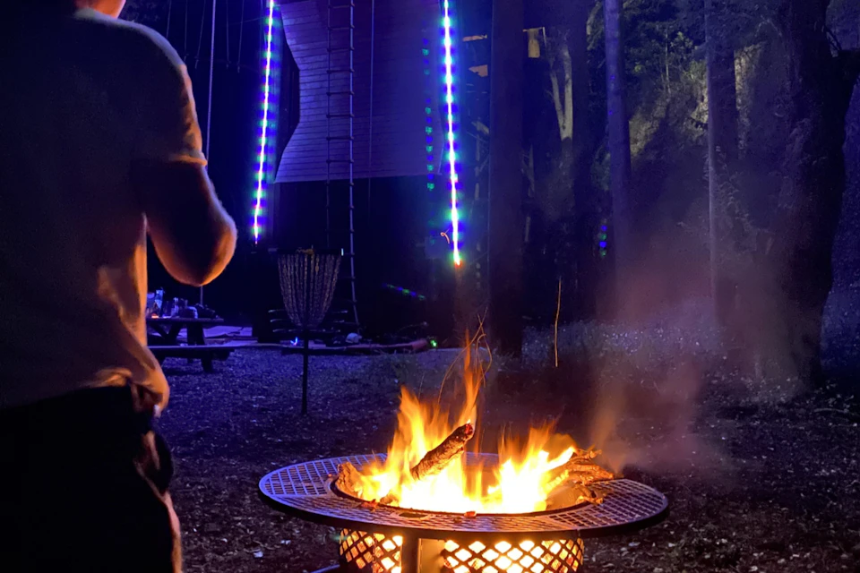 A campfire burns brightly in a metal fire pit at night, with a person standing nearby and colorful lights in the background.