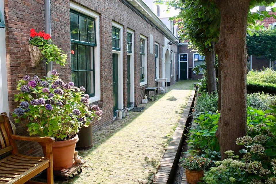 A narrow cobblestone alleyway lined with potted flowers and greenery, alongside brick buildings with green-trimmed windows and doors.