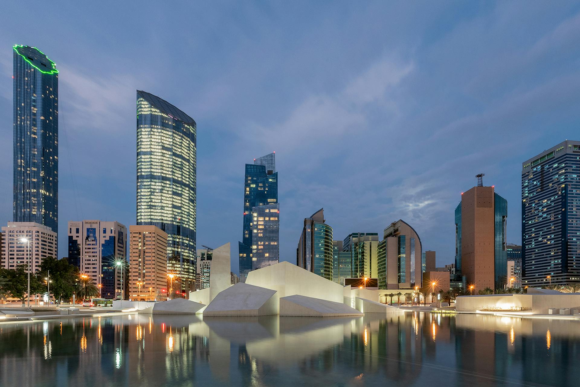 Modern city skyline at dusk with tall, illuminated buildings and unique white architectural structures reflected in a serene body of water.