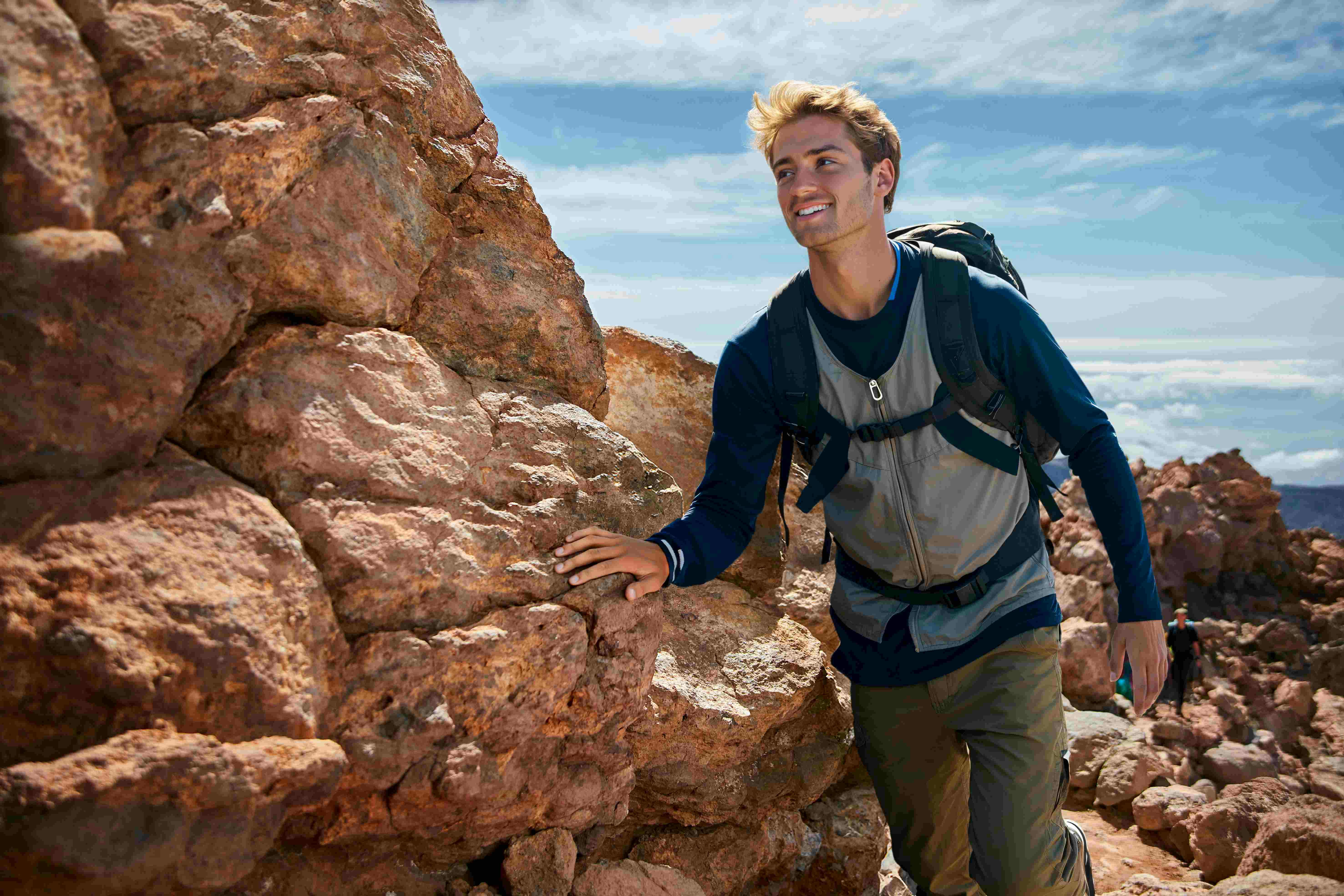 A man with a backpack climbs a rocky terrain under a partly cloudy sky, smiling as he navigates the rugged landscape.