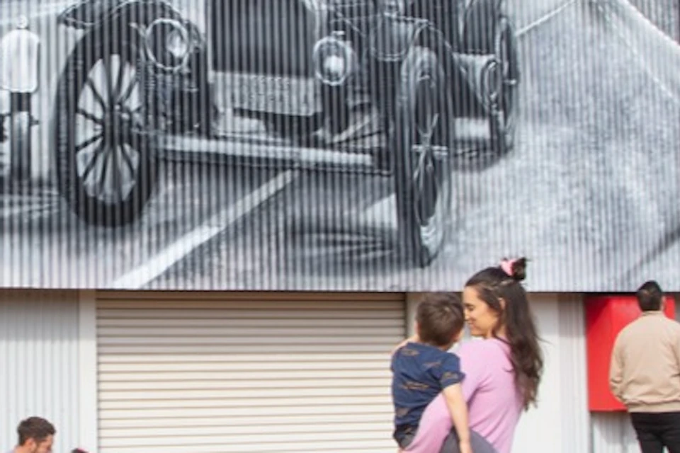 A woman holding a child stands in front of a building with a large mural depicting people and vintage cars. Several people sit nearby.