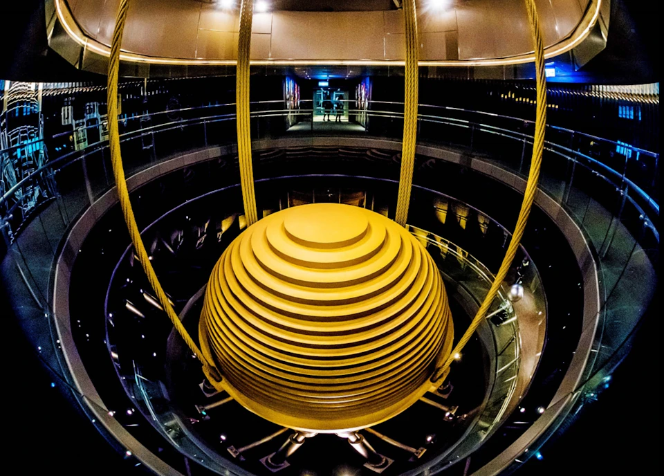 Large, yellow spherical structure suspended by cables in a circular, multi-level indoor space. Dim lighting.