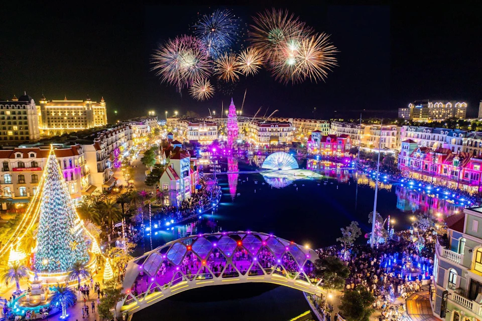 A vibrant nighttime festival with fireworks over a decorated cityscape, colorful lights, and a bridge over a water feature.
