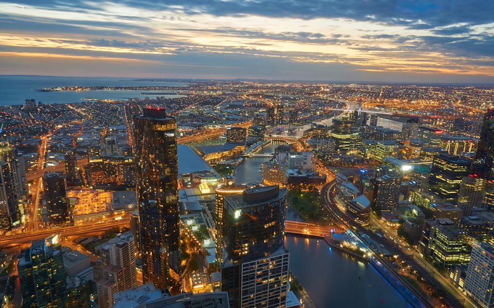 Aerial view of a cityscape at dusk with illuminated buildings, winding river, and distant horizon under a partly cloudy sky.