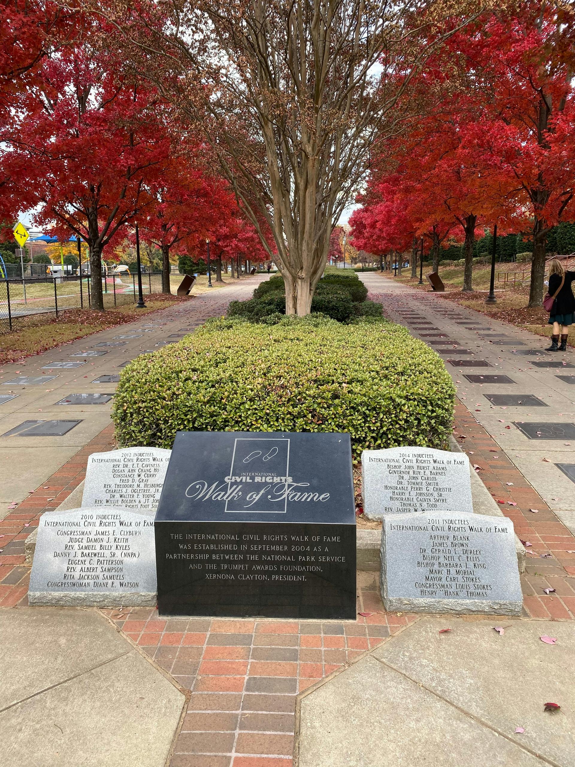 Allée bordée d'arbres à feuilles rouges, menant à une plaque noire "Civil Rights Walk of Fame" et à trois bornes en pierre. Deux personnes sont visibles au loin.