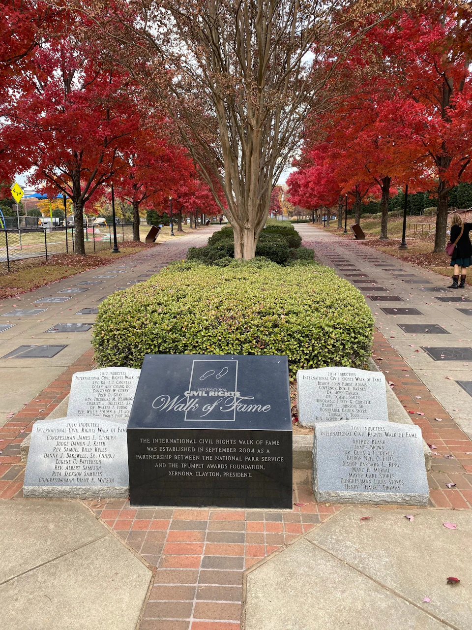 Pathway flanked by red-leaved trees, leading to a black "Civil Rights Walk of Fame" plaque and three stone markers. Two people visible in the distance.