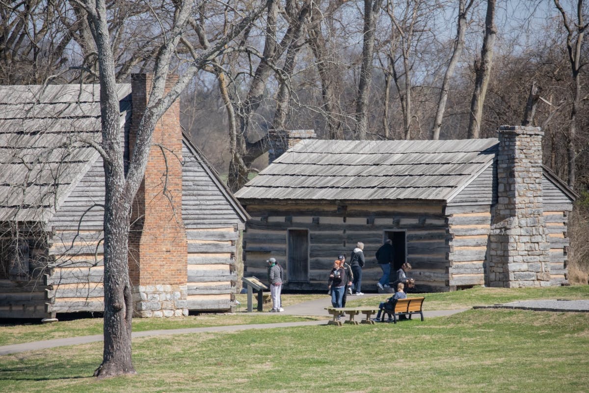 People exploring log cabins in a park with leafless trees in the background. A bench and a pathway are in the foreground.