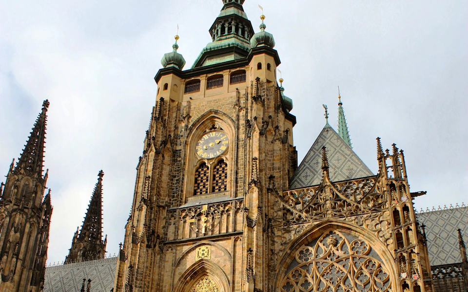 Ornate gothic cathedral facade with clock and detailed architectural carvings, set against a cloudy sky.