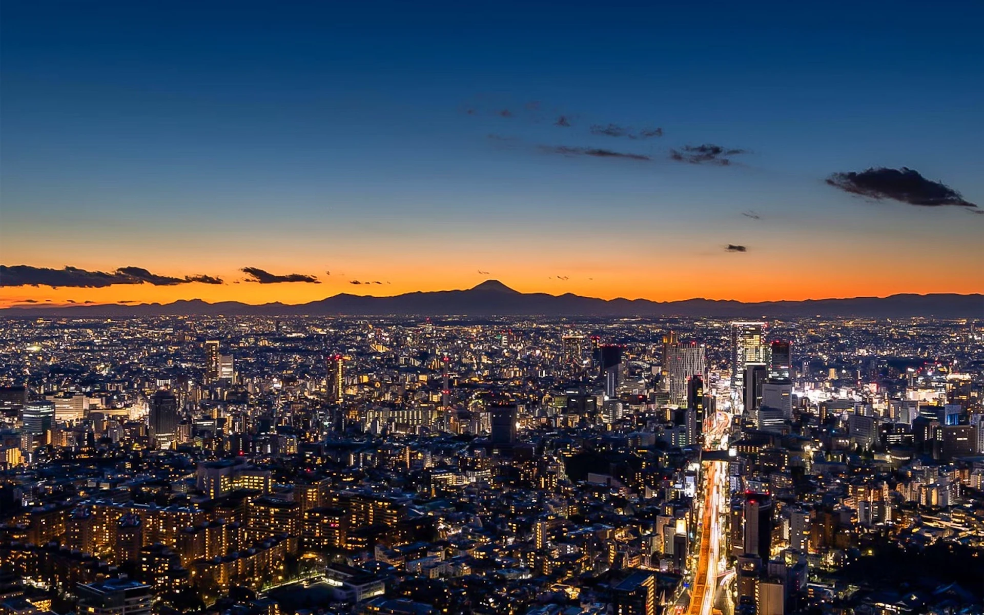 Cityscape at dusk with bright city lights and a silhouette of a mountain in the background against an orange sky.