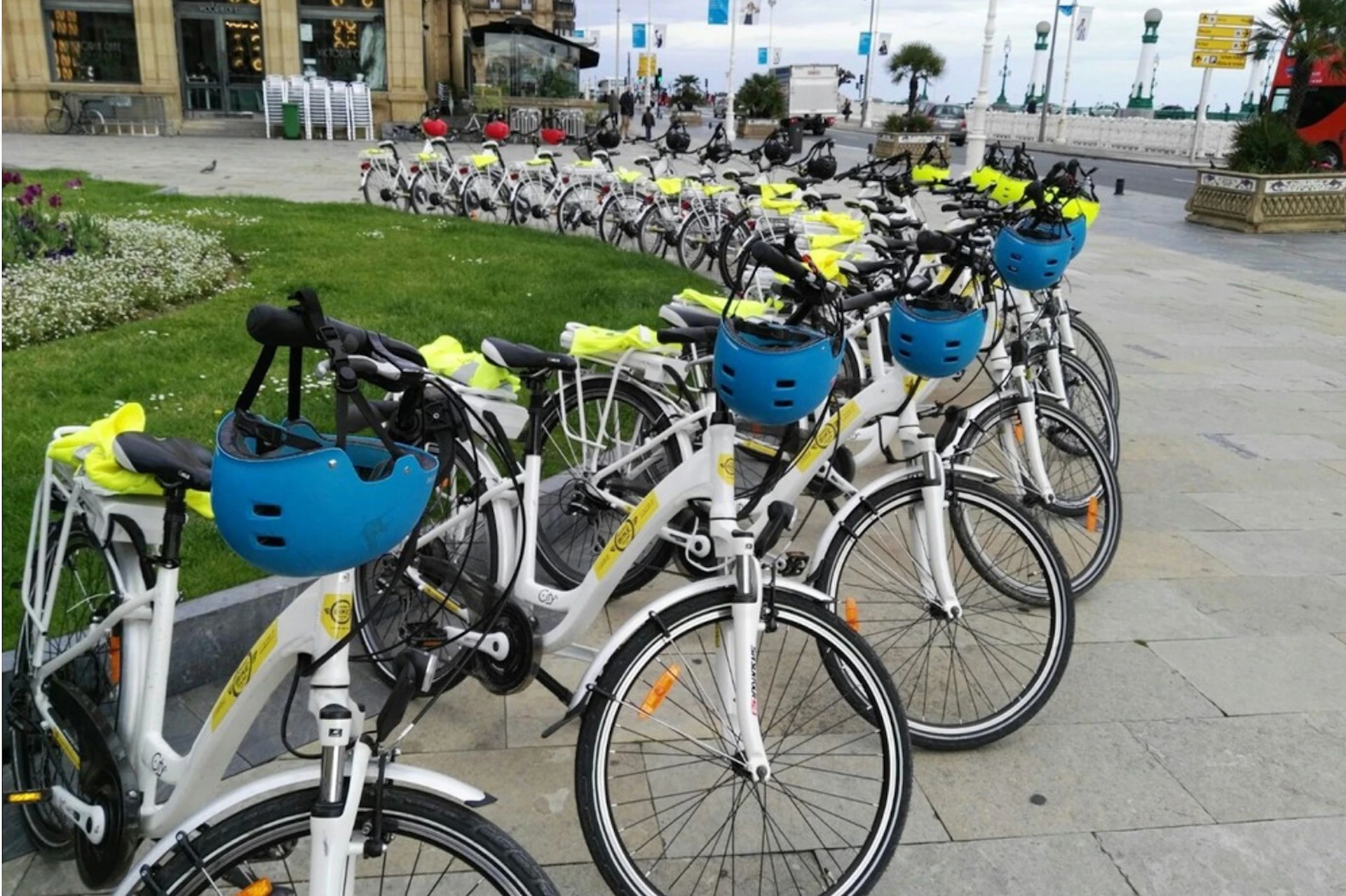Row of parked white rental bikes with blue helmets on handles, positioned along a city waterfront promenade.