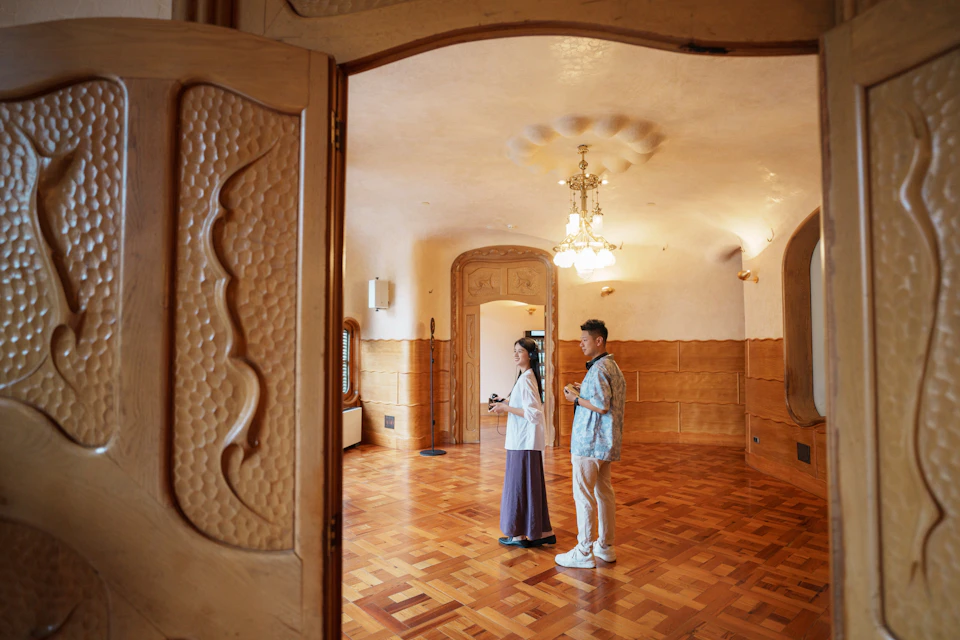 Two people stand in a wooden-floored room with ornate wooden doors and ceiling lights, viewed through a doorway.