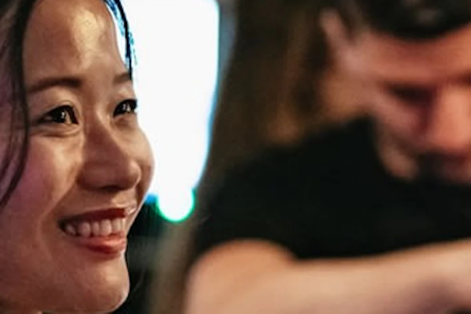 A woman smiles at a bar while a bartender pours drinks from a shaker in the background. Shelves of bottles are visible behind them.