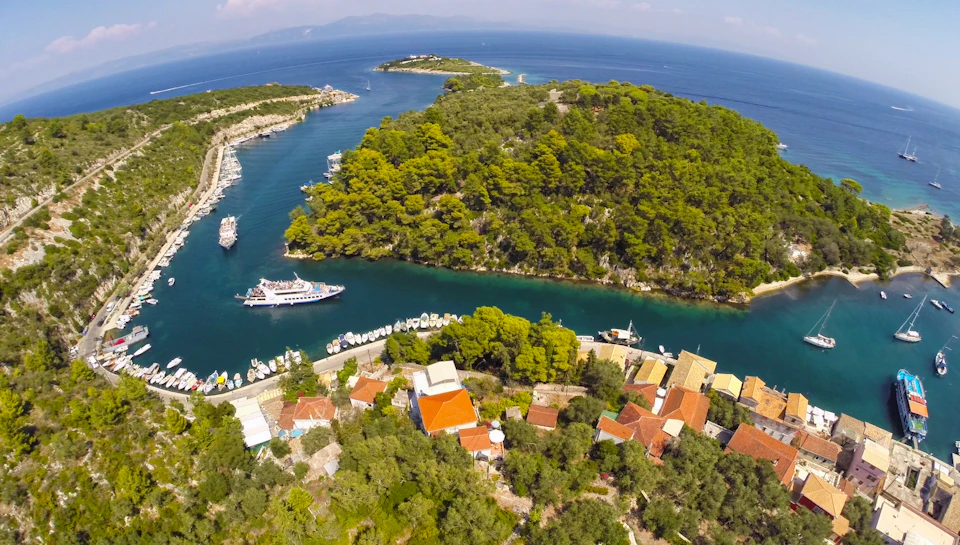 Aerial view of a coastal village with red-roofed houses, a dense forested peninsula, boats on a river, and surrounding blue sea.