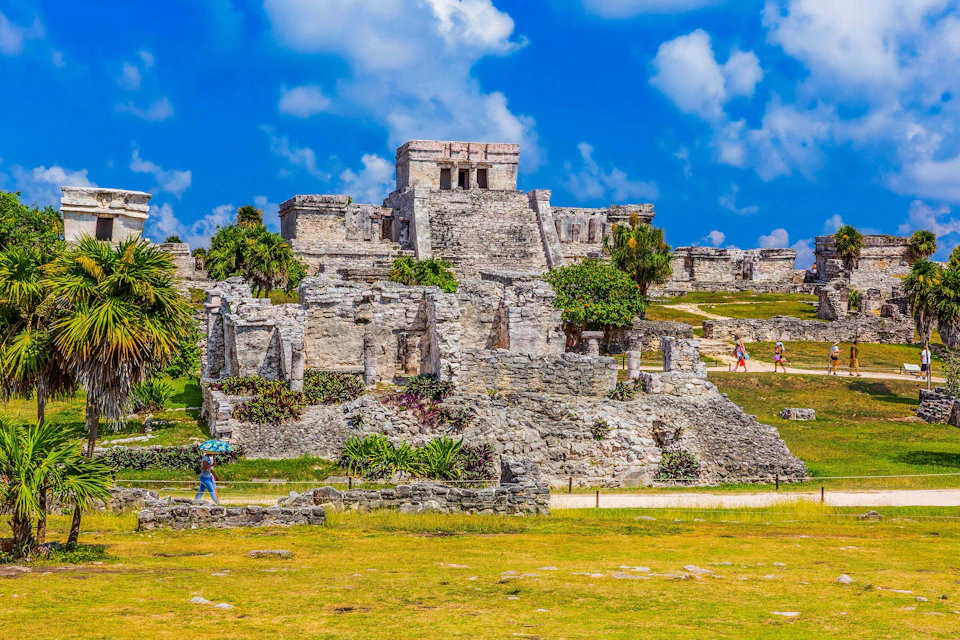 Ancient stone ruins with greenery under a partly cloudy sky, with a few people walking in the foreground.