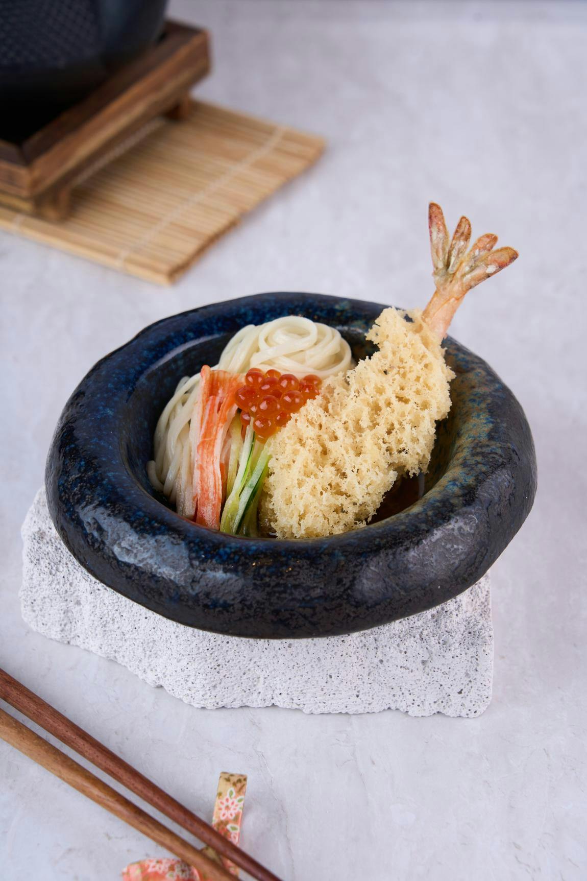 A bowl of noodles topped with tempura shrimp, fish roe, crab stick, and cucumber, served in a black ceramic bowl.