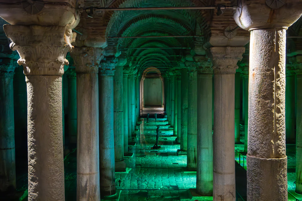 Ancient underground cistern with tall columns illuminated by green light, reflecting off shallow water on the floor.