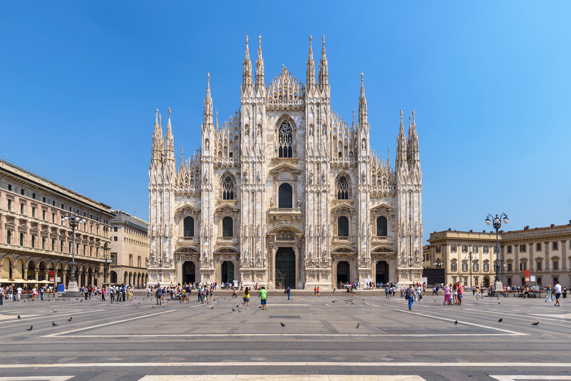 A large, ornate Gothic cathedral with ornate spires stands in a spacious plaza filled with people and pigeons under a clear blue sky.
