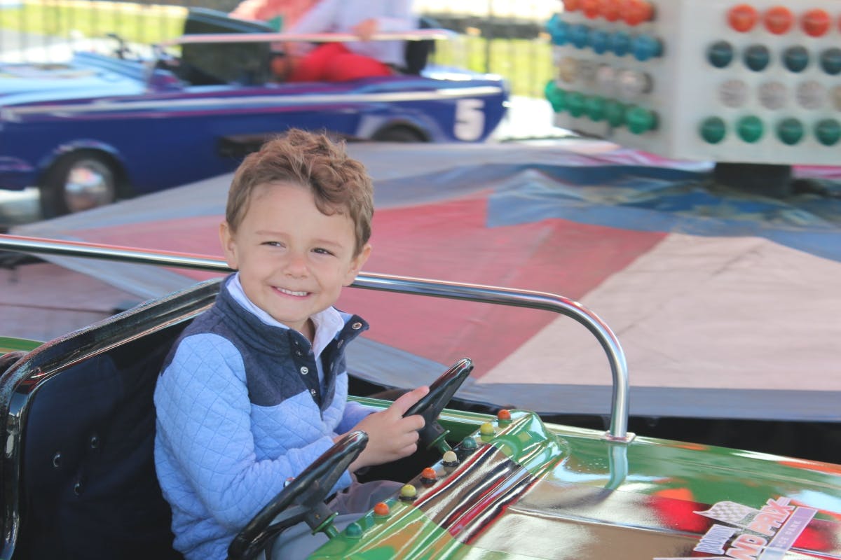 Young boy smiling while driving a green bumper car at an amusement park. Other bumper cars and colorful decorations are in the background.