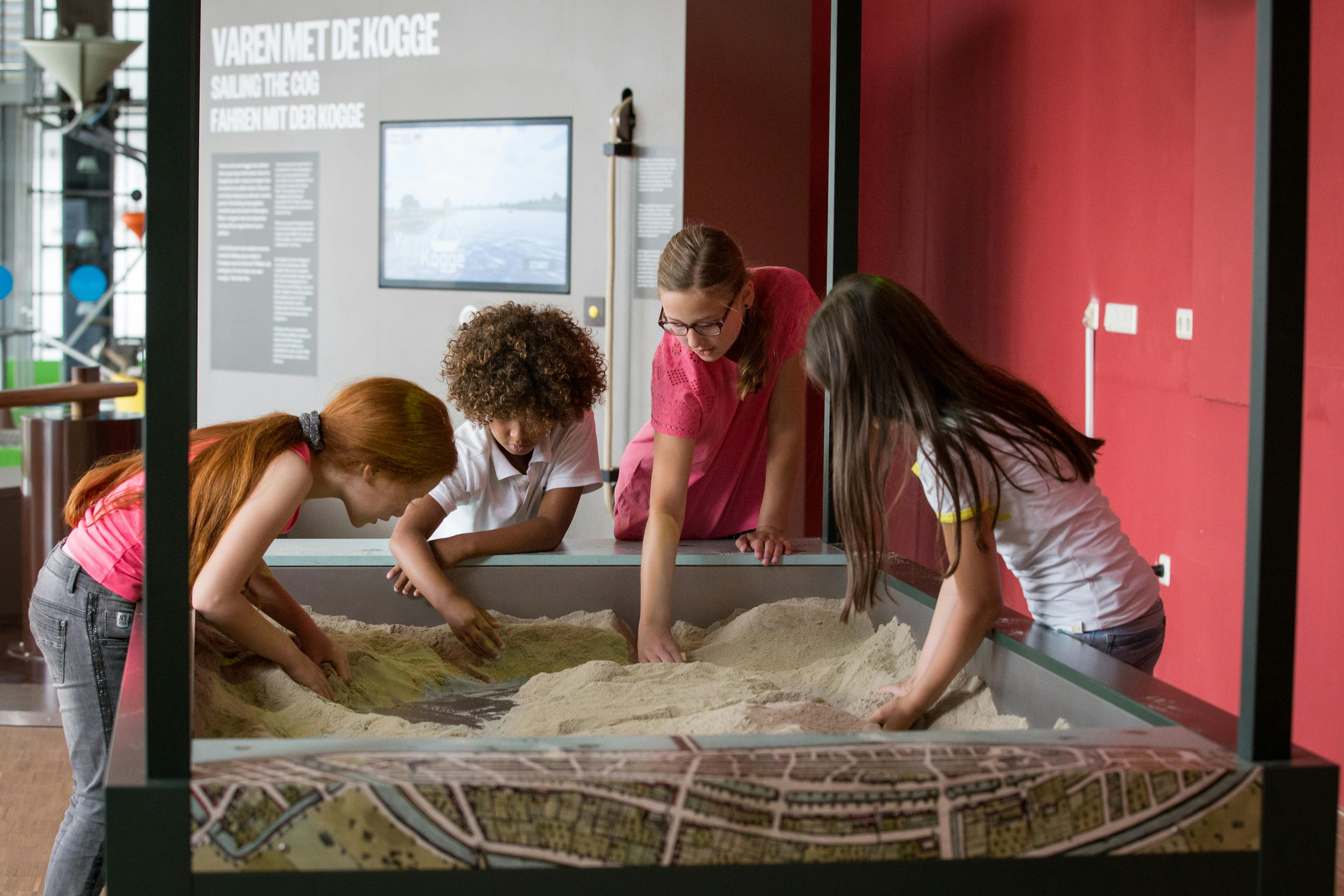 Čtyři děti si hrají s pískem ve velké interaktivní expozici v muzeu s informačními displeji na stěnách.