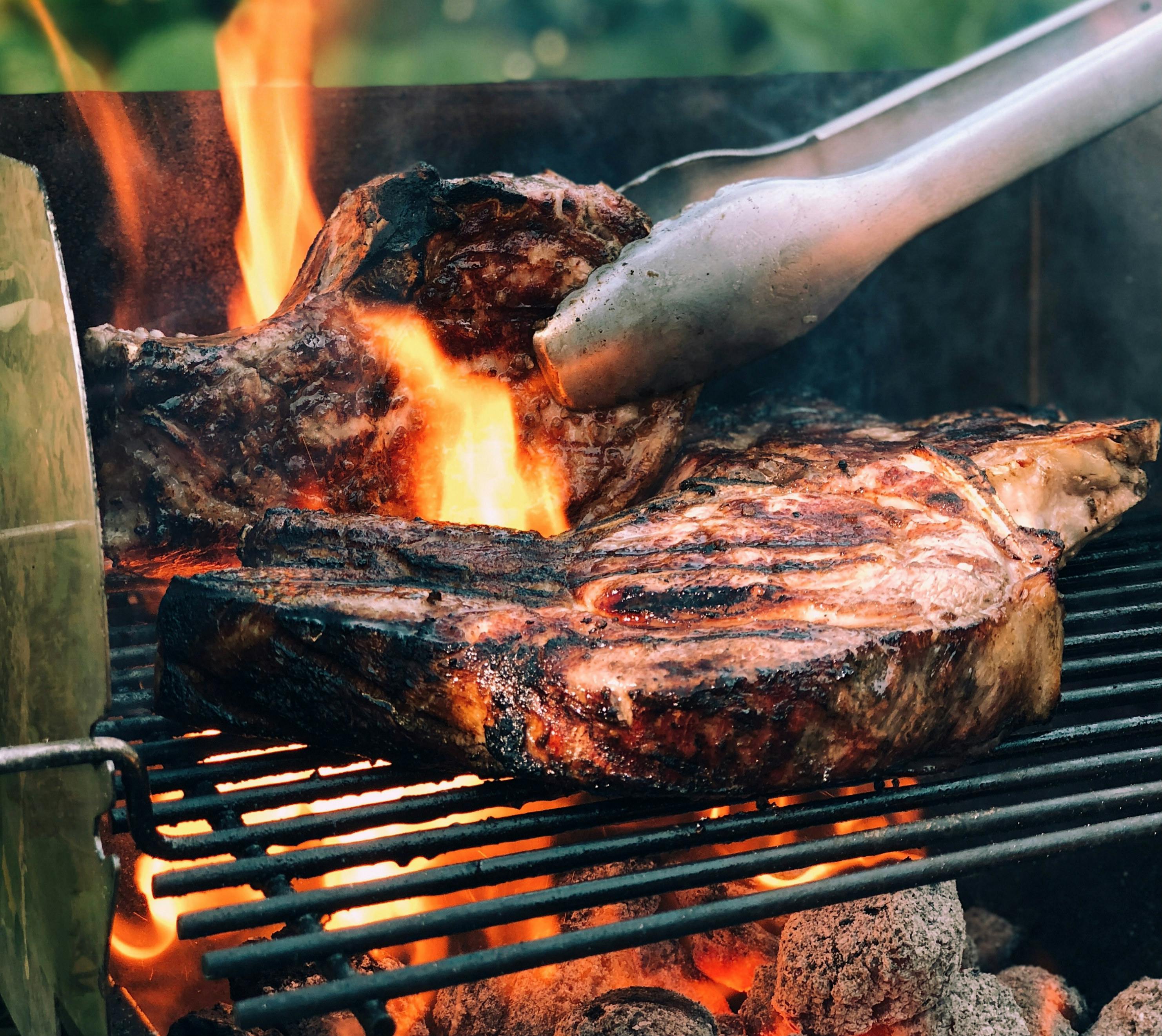 Close-up of large steaks grilling over an open flame, with metal tongs adjusting one steak on the grill.