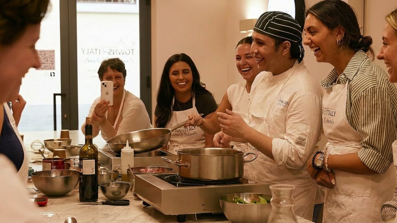 A group of people in a cooking class laughing around a kitchen counter with pans, pots, and ingredients.