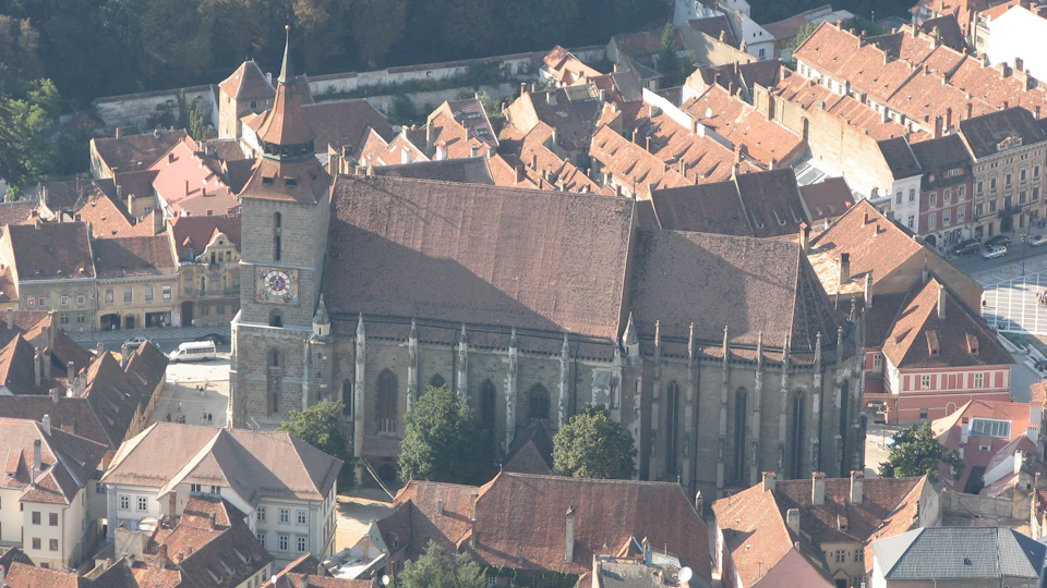 An aerial view of a large, historic church with a tall clock tower surrounded by tightly packed buildings with red-tiled roofs.