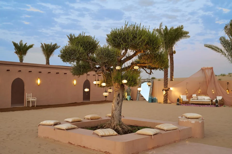 A courtyard with a tree decorated with lanterns, surrounded by a low wall with cushions, and palm trees in the background.