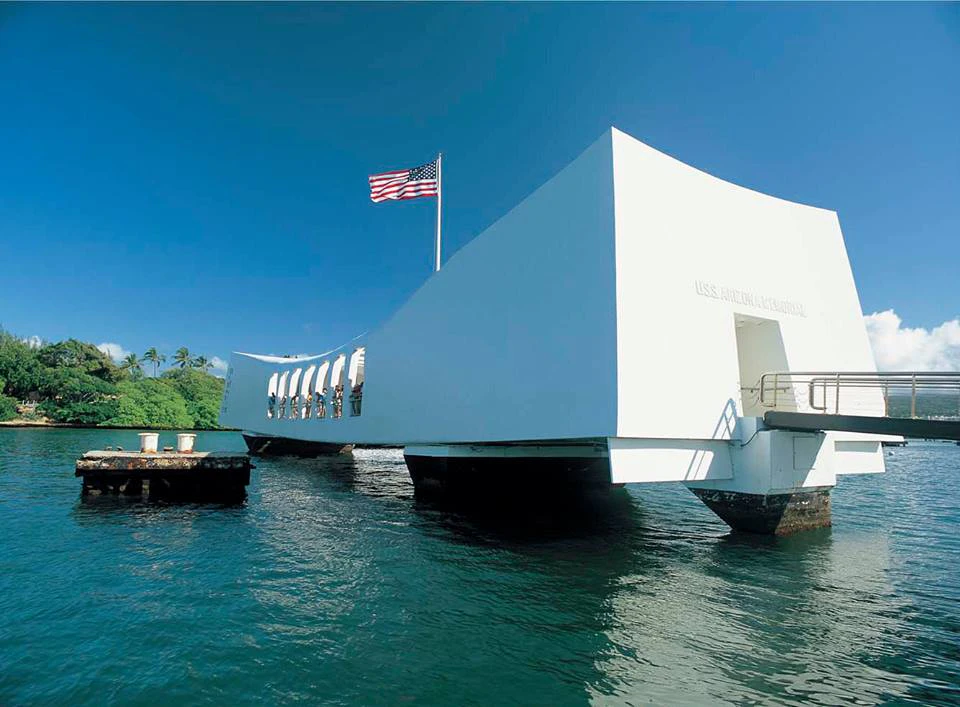 A white structure with an American flag stands over water under a clear blue sky, with greenery visible in the background.