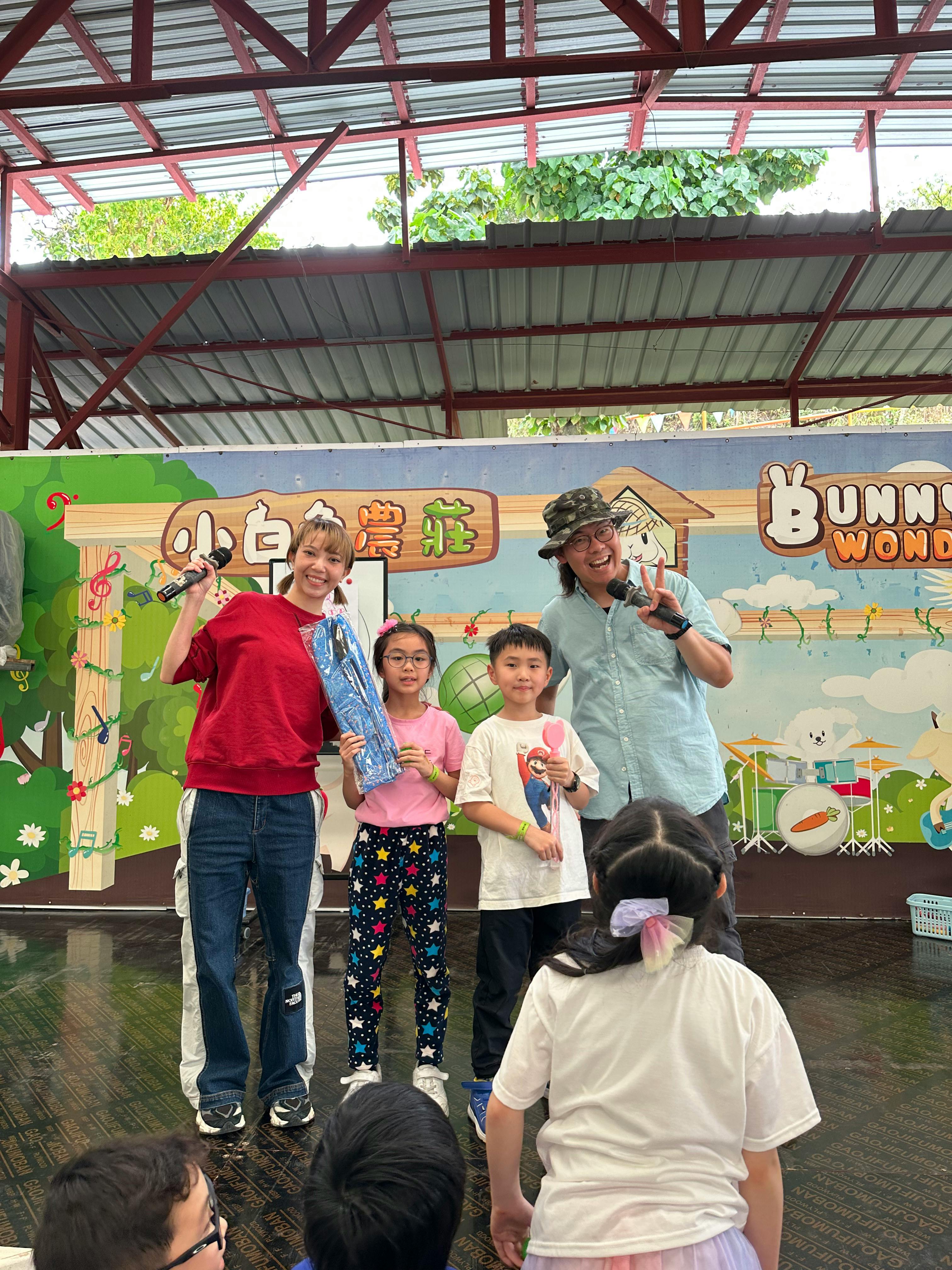 Two adults and two children stand smiling on a stage with a colorful background, while another child sits facing them.