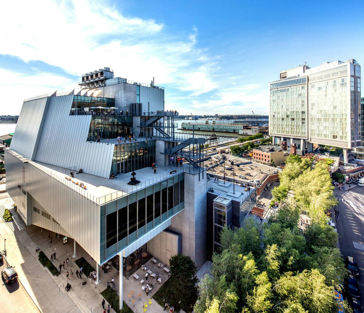 Un moderno edificio de varios niveles con terrazas y elementos acristalados, rodeado de árboles, junto a un paseo marítimo industrial y otro gran edificio.