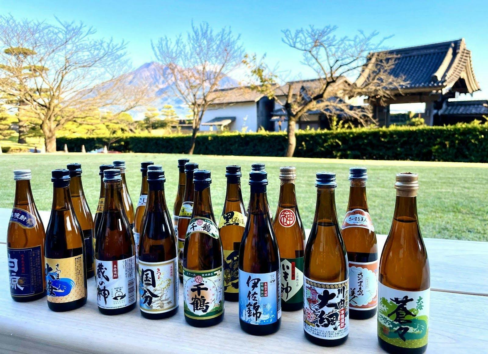 A variety of sake bottles are arranged on a table outdoors, with a traditional Japanese building and mountain in the background.