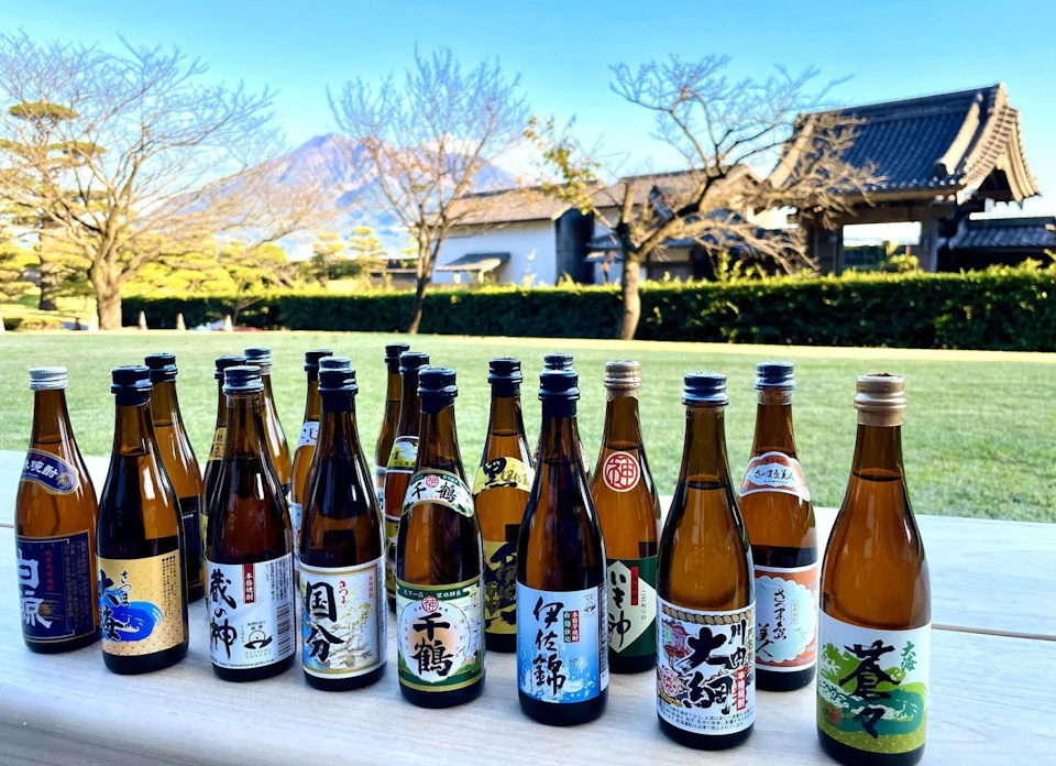 A collection of various labeled sake bottles displayed outdoors, with a traditional Japanese building and mountain in the background.