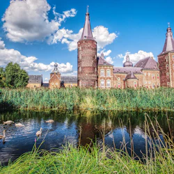 Historic brick castle with turrets, surrounded by greenery, adjacent to a reflective pond with swans. Bright blue sky with clouds.