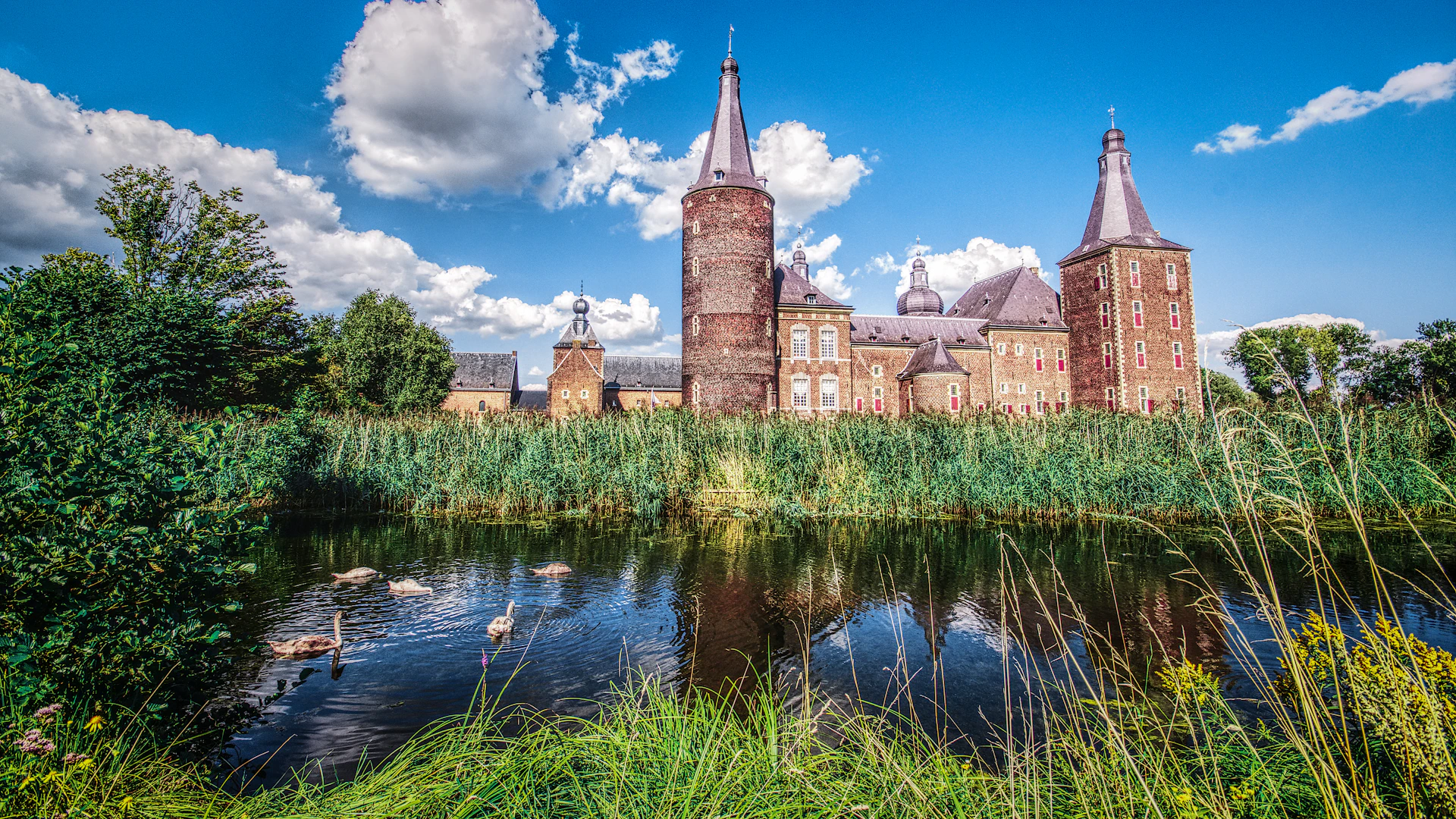 Historic brick castle with turrets, surrounded by greenery, adjacent to a reflective pond with swans. Bright blue sky with clouds.