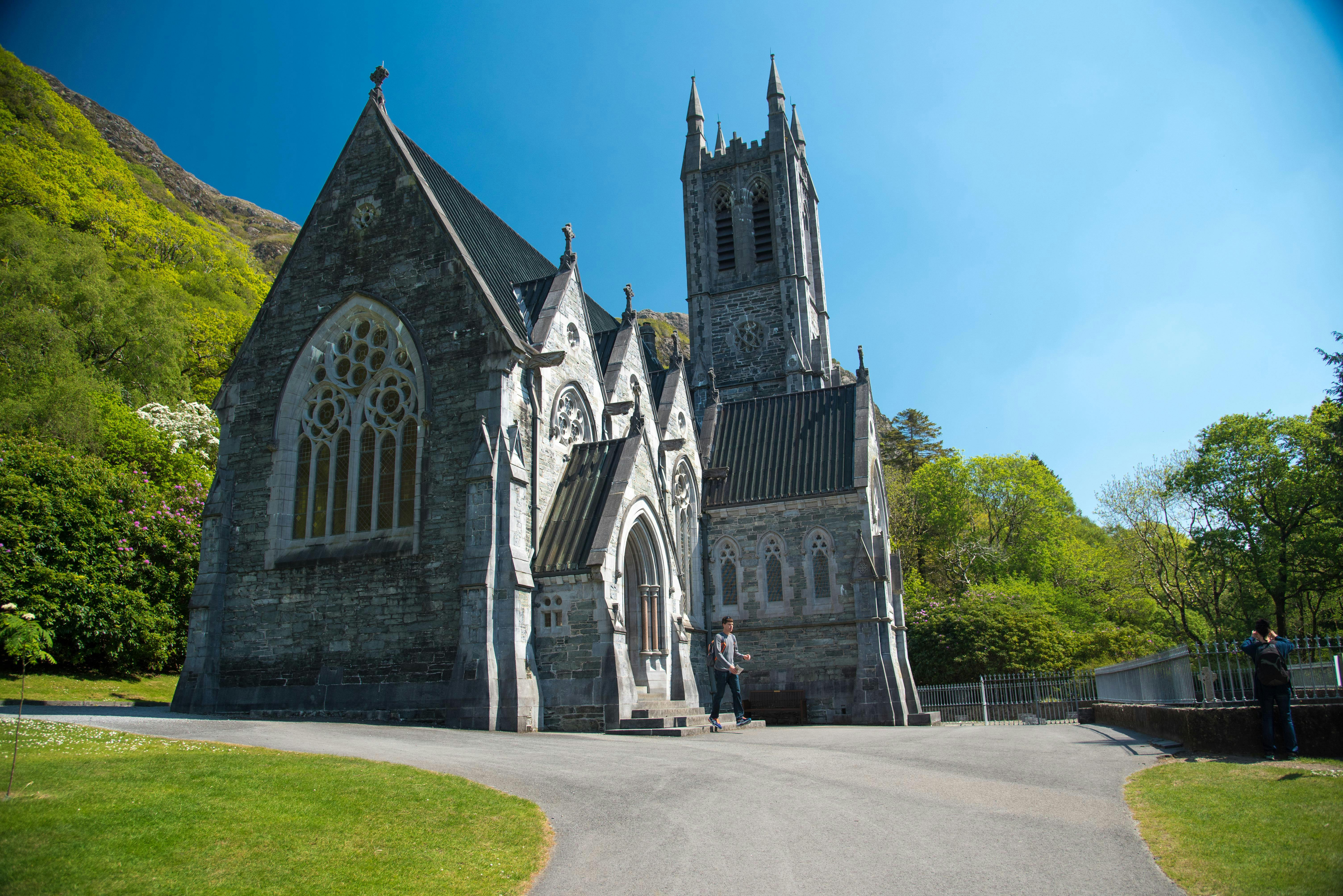 고딕 양식의 건축물, 스테인드글라스 창문, 첨탑, 시계, 아치형 입구가 있는 석조 교회. 나무로 둘러싸인 사람이 정면에 서 있습니다.