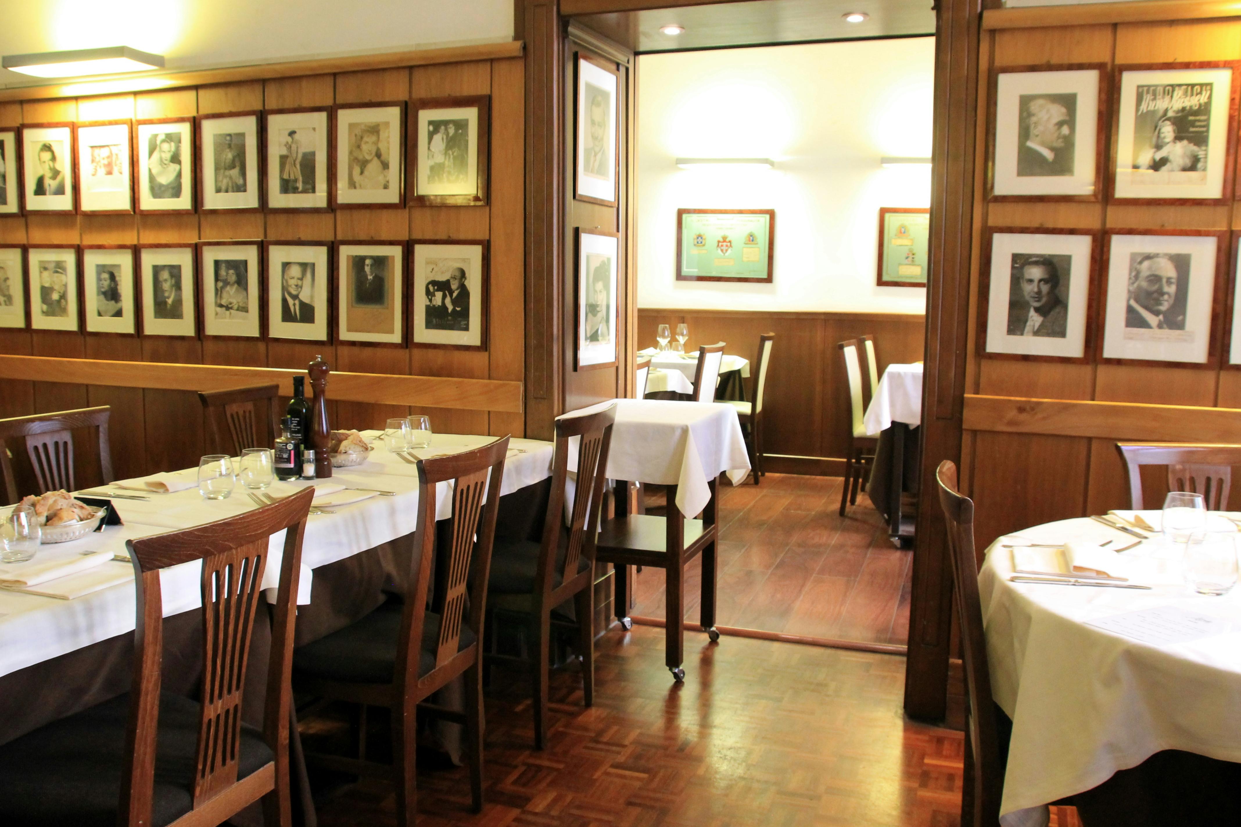 Уютный ресторан с белыми скатертями, деревянными стульями, обрамленными фотографиями на деревянных стенах и мягко освещенными обеденными зонами.