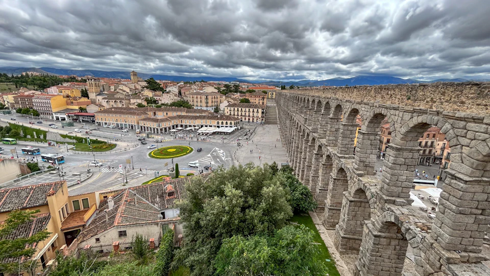 Ancient stone aqueduct in a historic town square, surrounded by buildings and greenery, under a cloudy sky.