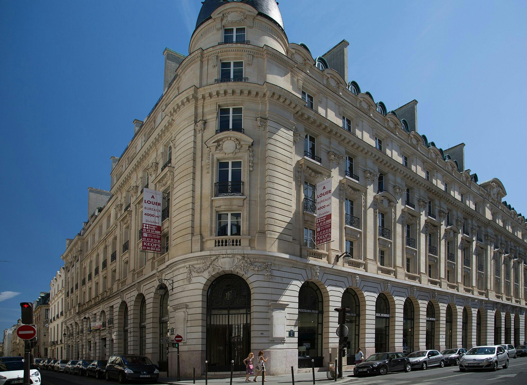 Angle d'un immeuble de plusieurs étages de style classique avec des panneaux de location "A Louer", des fenêtres cintrées et des voitures garées le long de la rue.