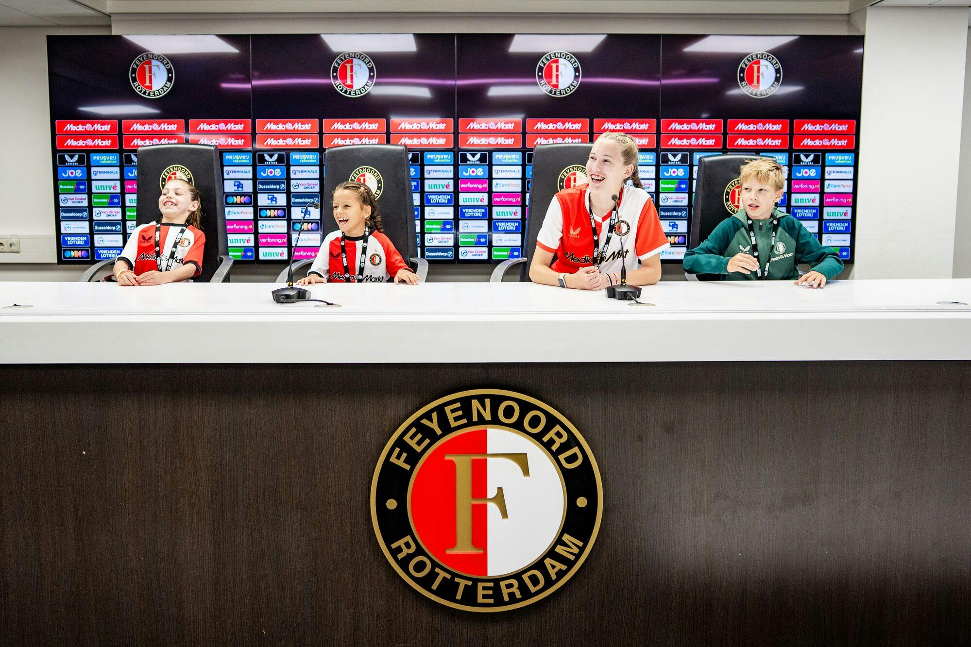 三个穿着Feyenoord球衣的孩子坐在新闻发布会上，带着麦克风，微笑着。
