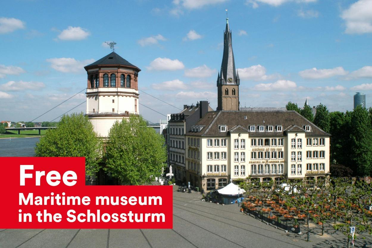 Ein Stadtplatz mit einem historischen Turm, einem Kirchturm und umliegenden Gebäuden unter einem blauen Himmel mit Wolken.