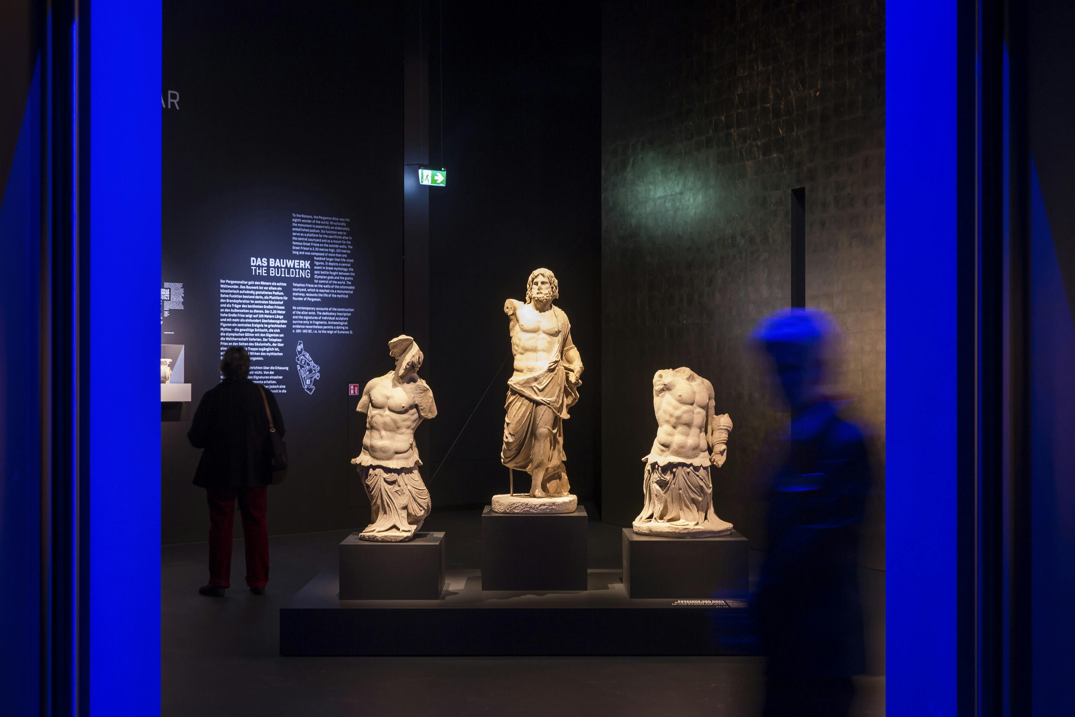 Ve slabě osvětlené muzejní expozici jsou vystaveny tři klasické sochy, dva fragmenty trupu a jedna celá postava. Návštěvníci se rozmazali v pohybu.