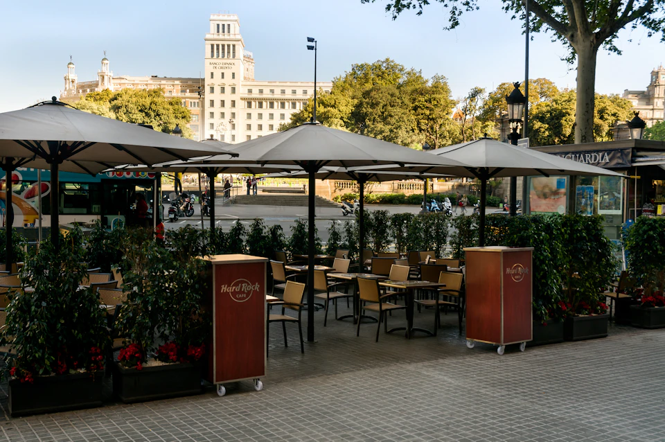 Outdoor café seating area with umbrellas and Hard Rock Café branding, trees in the background, and a large building visible behind.